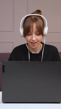 Uzaktan çalışan, kulaklık takan ve dizüstü bilgisayarda arka planda mor kanepe ve dikey videoda minimalist beyaz ofis bilgisayarında yazan mutlu, mutlu ve mutlu bir profesyonel.