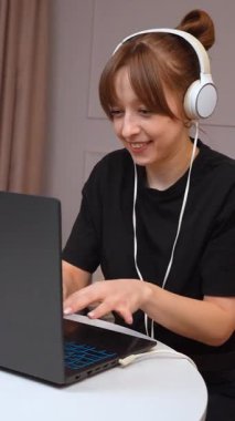 Uzaktan çalışan, kulaklık takan ve ev bilgisayarında yazı yazan genç bir kadın uzman dijital iş günü dikey videosu sırasında odaklanmış ve üretken davranışlar sergiliyor.