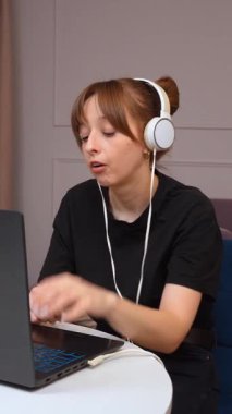 Uzaktan çalışan, kulaklık takan ve ev bilgisayarında yazı yazan genç bir kadın uzman dijital iş günü dikey videosu sırasında odaklanmış ve üretken davranışlar sergiliyor.