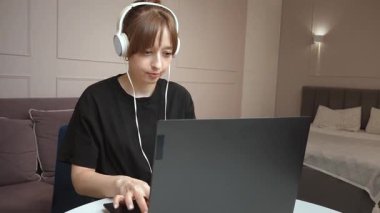 Uzaktan çalışan genç bir kadın, beyaz kulaklıklı dizüstü bilgisayar kullanırken rahat bir şekilde oturuyor ve sanal iletişim ve çevrimiçi buluşma ortamında profesyonelce iş görüyor.