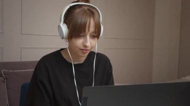 Uzaktan çalışan genç bir kadın, beyaz kulaklıklı dizüstü bilgisayar kullanırken rahat bir şekilde oturuyor ve sanal iletişim ve çevrimiçi buluşma ortamında profesyonelce iş görüyor.