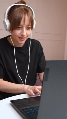 Uzaktan çalışan genç bir kadın, beyaz kulaklıklı laptop kullanırken rahat bir şekilde oturuyor ve sanal iletişim ve çevrimiçi buluşma ortamı dikey videosu ile profesyonelce bağlantı kuruyor.