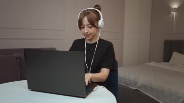 Uzaktan çalışan, dizüstü bilgisayarla yazarken dikkatle odaklanan, kulaklık takan ve rahat bir dairede, yuvarlak masada oturan serbest çalışan bir kadın.