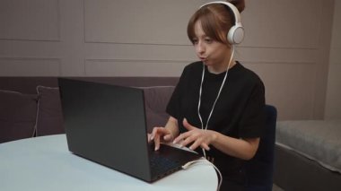 Evde çalışan, kulaklık takan ve bilgisayarındaki video konferansında konuşan genç iş kadını farklı ifadeler ve jestler sergiliyor.