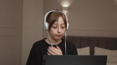 Evde çalışan, kulaklık takan ve bilgisayarındaki video konferansında konuşan genç iş kadını farklı ifadeler ve jestler sergiliyor.