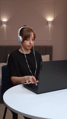 Evde çalışan, kulaklık takan ve bilgisayarındaki video konferansında konuşan genç iş kadını farklı ifadeler ve jestler gösteriyor.