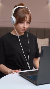 Evde çalışan, kulaklık takan, beyaz masada oturan ve dizüstü bilgisayara odaklanan, modern uzaktan kumandalı iş yaşam tarzı ve dijital verimlilik videolarını somutlaştıran kadın.