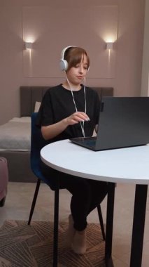 Evde çalışan, kulaklık takan, beyaz masada oturan ve dizüstü bilgisayara odaklanan, modern uzaktan kumandalı iş yaşam tarzı ve dijital verimlilik videolarını somutlaştıran kadın.