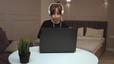 Evde çalışan, kulaklık takan, beyaz masada oturan ve dizüstü bilgisayara odaklanan, modern uzaktan kumandalı iş yaşam tarzı ve dijital verimliliği somutlaştıran bir kadın.