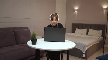 Evde çalışan, kulaklık takan, beyaz masada oturan ve dizüstü bilgisayara odaklanan, modern uzaktan kumandalı iş yaşam tarzı ve dijital verimliliği somutlaştıran bir kadın.