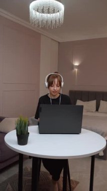 Evde çalışan, kulaklık takan, beyaz masada oturan ve dizüstü bilgisayara odaklanan, modern uzaktan kumandalı iş yaşam tarzı ve dijital verimlilik videolarını somutlaştıran kadın.