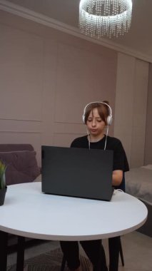 Evde çalışan, kulaklık takan, beyaz masada oturan ve dizüstü bilgisayara odaklanan, modern uzaktan kumandalı iş yaşam tarzı ve dijital verimlilik videolarını somutlaştıran kadın.