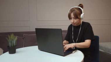 Uzaktan çalışan kulaklık takan genç profesyonel bir kadın, küçük yuvarlak masada otururken dizüstü bilgisayarda daktilo kullanıyor, rahat ofis ortamında dijital işlere odaklanıyor.