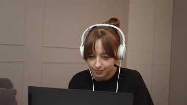 Uzaktan çalışan kulaklık takan genç profesyonel bir kadın, küçük yuvarlak masada otururken dizüstü bilgisayarda daktilo kullanıyor, rahat ofis ortamında dijital işlere odaklanıyor.