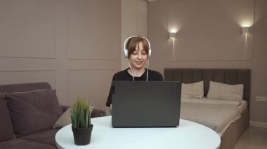 Uzaktan çalışan kulaklık takan genç profesyonel bir kadın, küçük yuvarlak masada otururken dizüstü bilgisayarda daktilo kullanıyor, rahat ofis ortamında dijital işlere odaklanıyor.