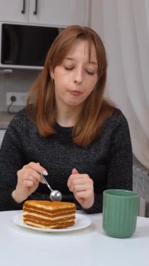 Milenyum kadını mutfak masasında oturmuş, katmanlı ballı kekin tadını çıkarıyor ve rahat, huzurlu bir ev ortamında, yumuşak, aydınlık, dikey video ile çaydan keyif alıyor.