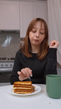 Sıcak içecekten ve tatlı tatlıdan hoşlanan genç bir kadın, modern mutfakta yumuşak doğal ışıkla oturuyor, rahatlamanın tadını çıkarıyor ve yemek pişirme zevkini dikey videoya alıyor.