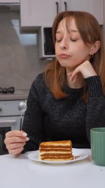 Katmanlı ballı pastanın tadını çıkaran genç bir kadın. Parlak, çağdaş beyaz bir mutfakta otururken kaşıkla narin ısırıkların tadını çıkaran bir kadın. Mutfak zevki ve kişisel rahatlama anı.