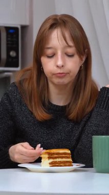 Katmanlı ballı pastanın tadını çıkaran genç bir kadın. Parlak, çağdaş beyaz bir mutfakta otururken kaşıkla narin ısırıkların tadını çıkaran bir kadın. Mutfak zevki ve kişisel rahatlama anı.