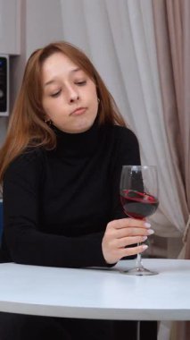 Modern bir mutfakta beyaz bir masada oturan genç bir kadın, sakin bir şekilde kırmızı şarabın tadını çıkarıyor, huzurlu bir rahatlama anı yaşıyor ve evde dikey videonun sessiz keyfine varıyor.