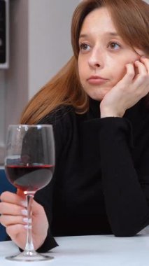 Modern bir mutfakta beyaz bir masada oturan genç bir kadın, sakin bir şekilde kırmızı şarabın tadını çıkarıyor, huzurlu bir rahatlama anı yaşıyor ve evde dikey videonun sessiz keyfine varıyor.