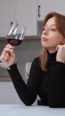 Beyaz mutfak masasında oturan kadın, sakin bir şekilde kırmızı şarabın tadını çıkarıyor, aromasını derin bir şekilde içine çekerken, aydınlık, çağdaş ev ortamında huzurlu bir aşçılık keyfi yaşıyor.