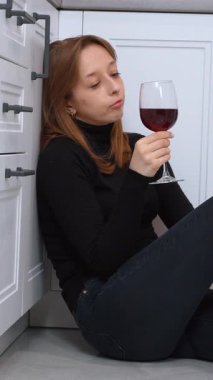 Duygusal olarak sıkıntılı genç bir kadın mutfakta oturmuş, yüz ifadelerini değiştirerek kırmızı şarap içiyor, derin kişisel mücadele ve iç karışıklığı dikey video çekiyor.