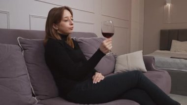 Oturma odasındaki rahat koltukta uzanan genç bir kadın, bir kadeh kırmızı şarabı nazikçe döndürüyor, sakin bir anı yaşıyor kişisel rahatlık ve sessiz bir ev keyfi