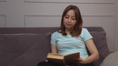 Kitap okuyan rahat bir kadın, bir dairenin oturma odasındaki koltukta rahat bir şekilde oturuyor, edebiyata dalmış, sessiz bir sessizlik ve rahatlık içinde.