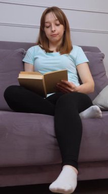 Rahat bir kadın kitap okuyor, bir dairenin oturma odasındaki koltukta rahat bir şekilde oturuyor, edebiyata dalıyor, sessiz bir sessizlik anında ve boş zamanlarında dikey bir videoda.