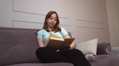 Bir kadın rahat bir şekilde kanepede oturup kitap okumaktan, duruşunu ve yüz ifadelerini değiştirmekten, sakin, huzurlu bir tavırla, sakin bir kişisel rahatlık anında, keyif almaktan hoşlanır.