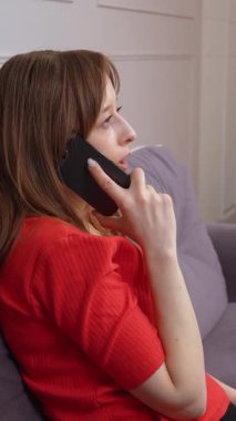 Kırmızı gömlekli genç bir kadın oturma odasındaki kanepede rahat bir şekilde otururken telefonda konuşuyor. Kişisel iletişim anındaki çeşitli duyguları dikey video görüntülerine yansıtıyor.