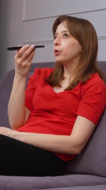 Genç kadın rahat bir şekilde oturma odasındaki koltukta oturuyor, modern akıllı telefonu kullanarak dijital sesli mesaj kaydediyor ve mobil teknoloji ile dikey video aracılığıyla iletişim kuruyor.