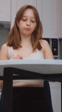 Genç bir kadın bir masada oturuyor, kaşıktan yemek tadıyor ve çeşitli düşünceli ve anlamlı yüz ifadeleri takınıyor, bir ev mutfağında çeşitli tepkiler ve duygular sergiliyor.