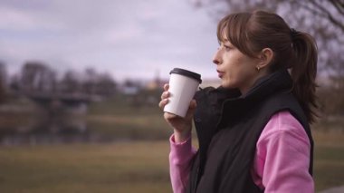 Açık havada, günlük kıyafetler giyen genç bir kadın akıllı telefonunu kullanırken kahve içmekten zevk alıyor, odaklanmaktan gülüşmeye kadar çeşitli ifadeler sergiliyor.