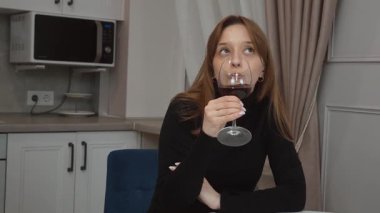 Modern bir mutfakta beyaz bir masada oturan genç bir kadın, sakin bir şekilde kırmızı şarabın tadını çıkarıyor, huzurlu bir rahatlama anı yaşıyor ve evde sakin bir akşam keyfi yaşıyor.