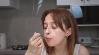 Mutfak masasında oturan genç bir kadın, yoğurttan hoşlanan, yüz ifadelerini değiştiren, özenli beslenen ve modern ev ortamında gündelik yaşam tarzı olan bir kadın.