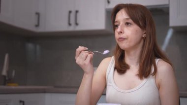 Genç bir kadın bir masada oturuyor, kaşıktan yemek tadıyor ve çeşitli düşünceli ve anlamlı yüz ifadeleri takınıyor, bir mutfak ortamında çeşitli tepkiler ve duygular sergiliyor.