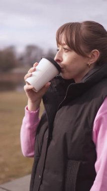 Genç bir kadın bankta oturuyor, kahve içiyor ve dinleniyor, açık hava, gözlük, şehir hayatı, açık hava kahvaltı konsepti dikey video