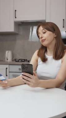 Mutfak masasında oturan genç bir kadın, mobil bankacılık uygulaması aracılığıyla güvenli bir şekilde çevrimiçi alışveriş yapıyor, kredi kartı tutuyor ve dijital satın almak için akıllı telefon kullanıyor.