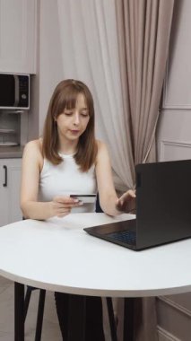 Genç profesyonel mutfak masasında rahatça oturuyor, elinde kredi kartıyla online alışveriş yapıyor, bilgisayarla dijital işlemleri dikey konsantrasyon videosuyla yönetiyor.