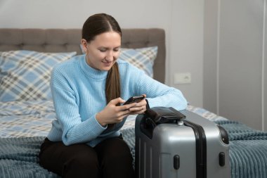 Genç bir kadın bavulunun yanında bir yatakta oturuyor, akıllı telefonunu kullanarak yaklaşan bir iş gezisi ya da tatil için seyahat planlarını tamamlıyor, modern teknolojiyi kolaylık için kucaklıyor.