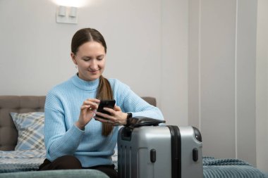 Akıllı telefon kullanan genç bir kadın valizinin yanında dinlenirken iş seyahatine ya da tatile çıkmaya hazır seyahat planlama ve internet rezervasyonunun keyfini çıkarıyor.