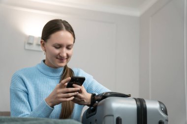 Akıllı telefon kullanan genç bir kadın bavulunun yanında oturuyor, seyahat planlıyor ve online rezervasyon yaptırıyor ya da seyahat bilgilerini kontrol ediyor.