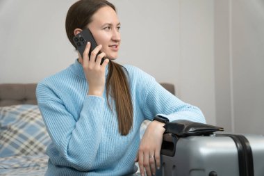 Mavi kazak giyen genç bir kadın, bavuluna yaslanırken bir telefon görüşmesi yapıyor. Yolculuğa hazır, yatak odası düzeninde, her an ayrılabileceğini gösteriyor.