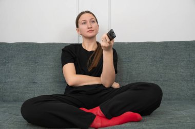 Gri bir kanepede bacak bacak üstüne atmış, elinde uzaktan kumandayla oturan rahat bir kadın, en sevdiği TV programını eğlence ve rahatlamaya dalmış bir oturma odasında keyifle izliyor.