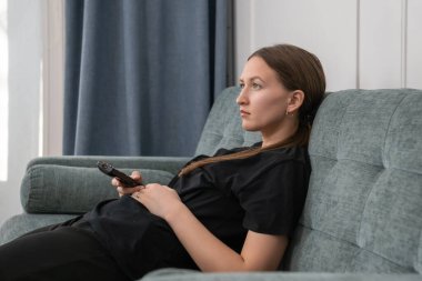 Sıkıcı genç kadın rahat bir kanepede uzanıyor, elinde uzaktan kumandayla oturma odasında televizyon seyrediyor, gün boyunca pasif rahatlık ve rahatlık içinde.