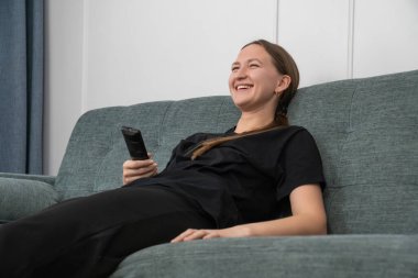 Mutlu genç kadın rahat bir kanepeye uzanıyor, uzaktan kumandayı tutuyor, komedi filmi veya TV programı izliyor, gülüyor ve eğleniyor, evde boş zamanlarının tadını çıkarıyor.