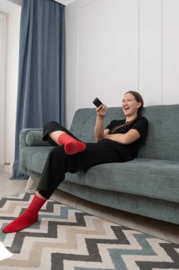 Kırmızı çorap giyen genç kadın oturma odasında rahat bir koltukta oturuyor, uzaktan kumandayı tutuyor ve televizyonda komedi programı izlerken gülüyor. Dikey fotoğraf