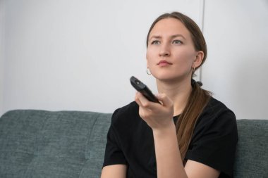 Oturma odasındaki rahat bir koltukta oturan, elinde uzaktan kumandayla televizyon izleyen, evinde eğlenen genç bir kadın.